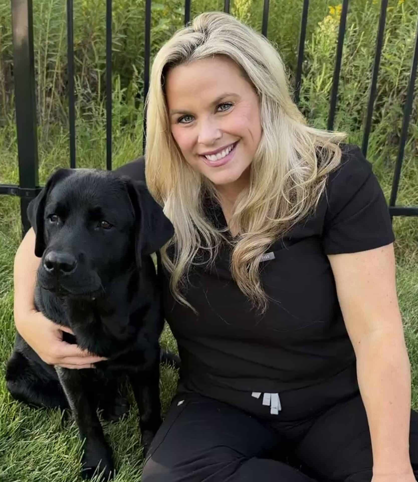 Woman with long blonde hair wearing black clothing sits on grass next to a black Labrador Retriever, smiling at the camera. Black metal fence and greenery are in the background.