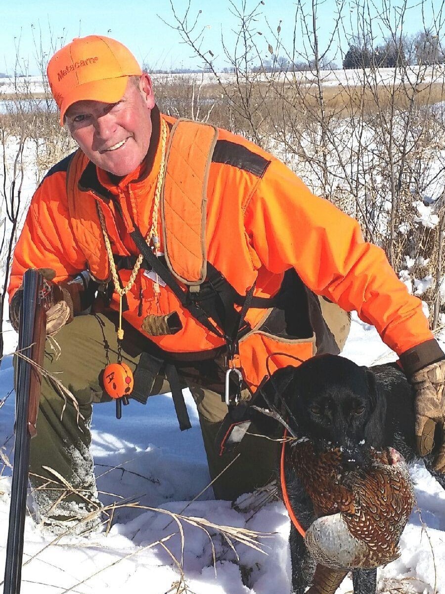 A hunter in blaze orange gear kneeling in snow with a black dog carrying a pheasant in its mouth.